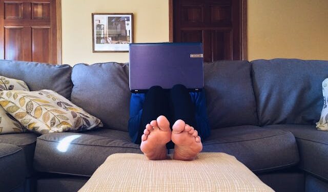 Person on a sofa with face hidden behind a laptop; only their feet are visible. Working at home.